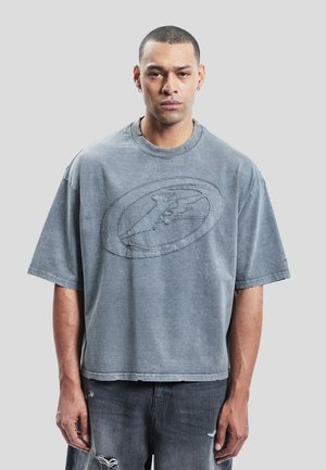 Mann med kort hår og øredobber iført en løs, falmet grå T-skjorte med et sydd ovalt mønster og revne mørke jeans.