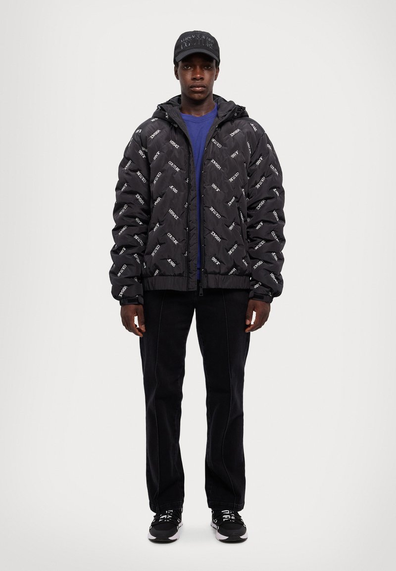 Chaqueta tipo puff en negro con impresiones de logotipo en blanco, combinada con una camisa azul, pantalones negros y zapatillas negras. Incluye una gorra a juego.