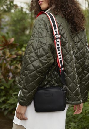Mujer con chaqueta acolchada verde lleva un bolso bandolera negro con una correa ancha etiquetada "HUNTER" en rojo, blanco y negro.