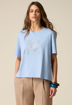 Junge Frau trägt hellblaues T-Shirt mit Strassstein-Hundedesign und beige plissierte Hose, posiert mit blauen und schwarzen Armreifen.