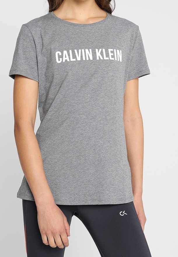Femme portant un t-shirt gris à manches courtes Calvin Klein et des leggings noirs avec un petit logo CK sur la cuisse.