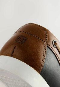 Chaussure en cuir marron avec une section noire, matériau texturé, semelle en caoutchouc blanc et détails de couture visibles. Logo embossé à l'arrière.