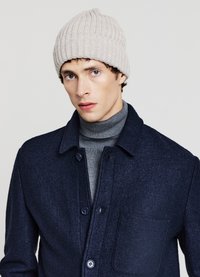 Cappello beige a costine indossato da una modella, abbinato a una giacca camicia in lana blu scuro e a un maglione dolcevita grigio aderente.