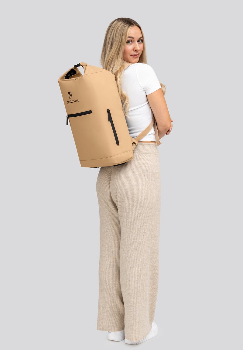 Mochila impermeable beige con un diseño de rollo en la parte superior, bolsillo con cremallera negra y correas ajustables, llevada en la espalda sobre un fondo gris.