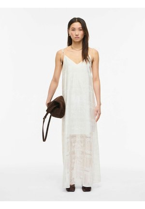 Mujer que lleva un vestido largo blanco sin mangas con detalles de encaje, sosteniendo un bolso de ante marrón y con zapatos oscuros, de pie frente a un fondo blanco.