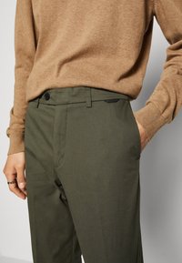 Olivgröna byxor med raka ben, med knappstängning, sidofickor och en slät textur. Bärs med en ljusbrun stickad tröja.