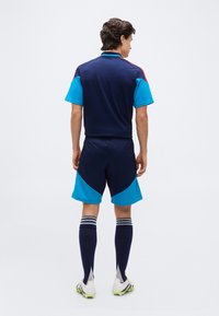 Man wearing navy and bright blue sportswear, striped knee-high socks, and white cleats, standing facing away on plain background.