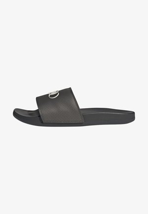 Svart slidesandal med perforerad ovandel, robust flat sula och en rund metallaccent på remmen. Mjuk textur överallt.