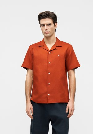 Homme portant une chemise à manches courtes orange brûlé avec un petit logo sur la poitrine gauche, associée à un pantalon foncé, debout devant un fond uni.