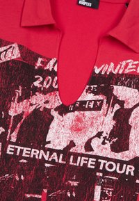 Chemise en coton rouge avec un imprimé graphique en noir et blanc, incluant du texte qui dit "TOUR DE LA VIE ÉTERNELLE". Col en V.