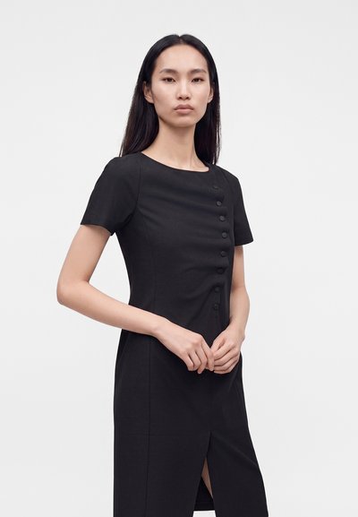 Femme aux longs cheveux foncés portant une robe noire à manches courtes avec des boutons sur le devant, debout devant un fond clair uni.
