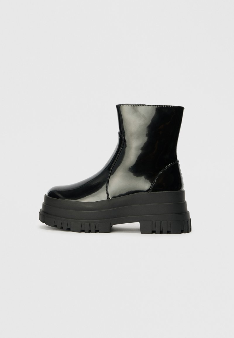 Must mustakate läikiv ankle boot paksu lõkkeka üle ja ümarate varvastega, külgvaates halli ühtlase taustal.