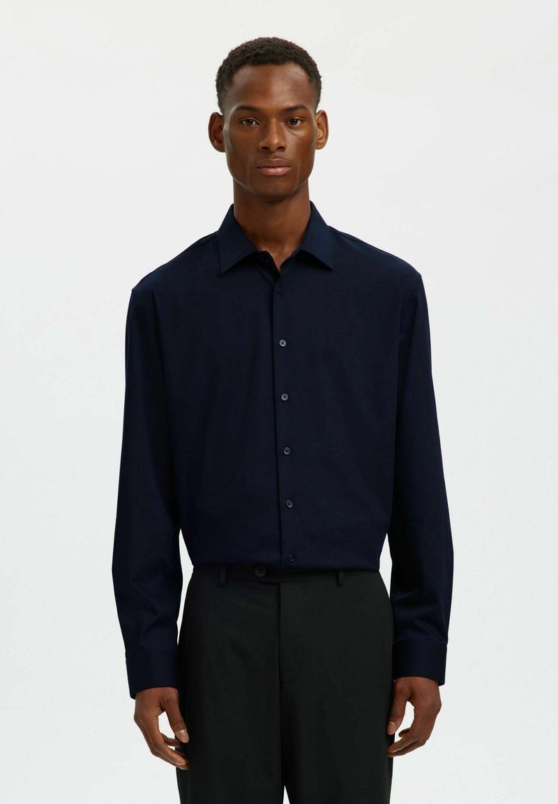 Chemise bleu marine à manches longues, boutonnée, avec un col classique, une texture lisse et des poignets à boutons, associée à un pantalon tailleur foncé.