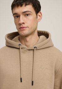 Beżowa bluza z kapturem z sznurkiem o czarnych końcach i haftowanym logo na lewej piersi. Miękka tkanina o luźnym kroju i kieszeni kangurzej.