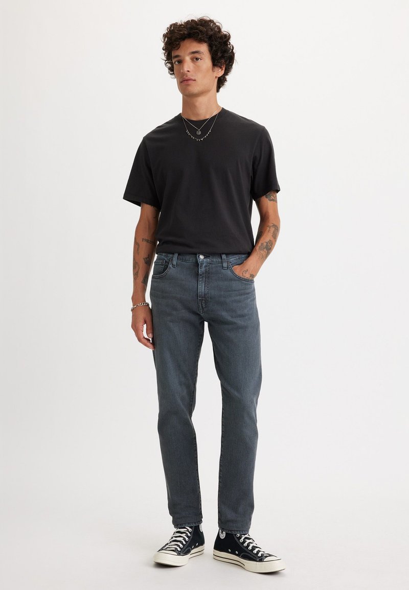 Schwarzes T-Shirt mit kurzen Ärmeln, graue Jeans und schwarze High-Top-Sneaker. Das Modell steht mit den Händen in den Taschen vor einem weißen Hintergrund.