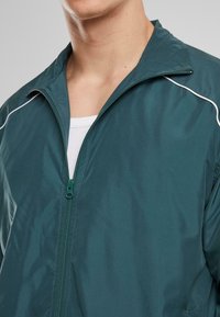 Menns zip-up jakke i mørkegrønn, med lyse kanter langs sømmene, laget av glatt, lett materiale med stående krage.