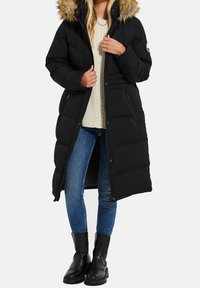 Femme portant un long manteau noir matelassé avec capuche en fourrure, pull crème, jean bleu et bottes noires, debout devant un fond blanc.