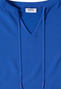Vêtement bleu avec col en V et cordons, tissu texturé et étiquette blanche cousue à l’intérieur du col.