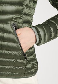 Green puffer jacket with quilted texture, featuring a zippered pocket and soft cuffs. Material appears smooth and slightly glossy.