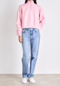 Pullover in pile rosa con collo alto e zip, vita elastica; abbinato a jeans azzurri e sneakers bianche. Texture liscia su tutta la superficie.