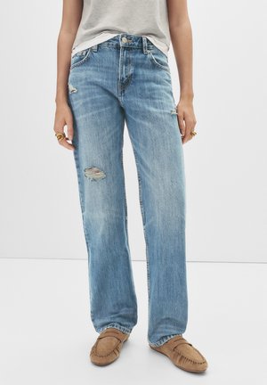 Light blue straight-leg jeans with distressed holes, worn with beige moccasin shoes and a tucked-in light gray sleeveless top.
