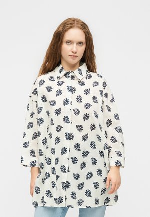 Vrouw met lang rood haar draagt een wit oversized blouse met knopen en een patroon van zwarte bladeren en lichtblauwe spijkerbroek.