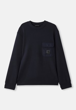 Un sweatshirt bleu marine en tissu doux, avec une poche frontale et un patch carré. L'encolure ronde et les poignets côtelés complètent le design.