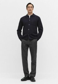 Marinblå ullskjorta med knappar och krage, framficka och blå knappar, matchad med grå texturerade byxor och svarta loafers.