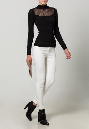 Donna che indossa un top nero a maniche lunghe con scollatura in rete, pantaloni skinny bianchi, stivaletti neri con tacco e tiene in mano una clutch beige.