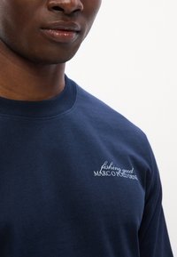 Tumesinine puuvillane särk, millel on ümmargune kaelus, ribitud krae ja valge tikandiga tekst detailid rinnal.