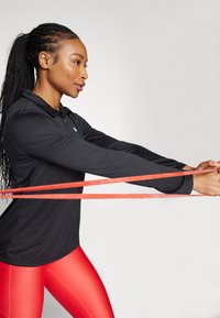 Haut de sport à manches longues noir et leggings rouges brillants. Le sujet tient des bandes de résistance rouges, mettant en valeur l'ajustement et la flexibilité des vêtements.