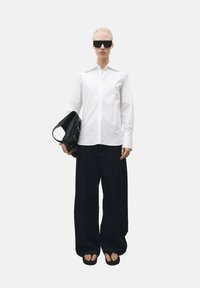 Weißes Hemd mit hohem Kragen, schwarze weitgeschnittene Jeans, große schwarze Sonnenbrille und eine schwarze Handtasche in der linken Hand.