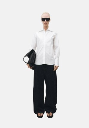 Weißes Hemd mit hohem Kragen, schwarze weitgeschnittene Jeans, große schwarze Sonnenbrille und eine schwarze Handtasche in der linken Hand.