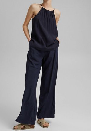 Femme portant un haut dos nu bleu marine ample, un pantalon large assorti et des sandales beiges à brides croisées, debout les mains dans les poches devant un fond gris.