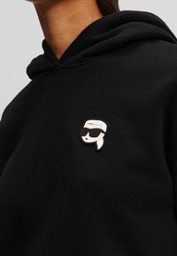 Czarna bluza z kapturem z gumową przypinką przedstawiającą stylizowaną postać z białymi włosami i okularami przeciwsłonecznymi, umieszczoną na piersi.