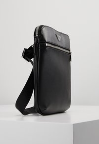 Borsa a tracolla nera in materiale sintetico con superficie testurizzata, chiusura a zip, tracolla regolabile e un piccolo logo sul fronte.