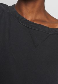 Musta collegepaita, jossa leveä pääntie, teksturoitu kangas ja näkyvät ompelusaumat kauluksen ja hartiasaumojen ympärillä.