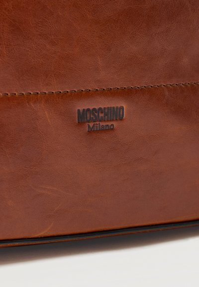 Surface en cuir marron avec des coutures visibles et un logo embossé "Moschino Milano" en lettres foncées.