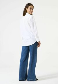 Vrouw in een wit shirt met lange mouwen en een wijde spijkerbroek, die over haar schouder kijkt, staande op een witte achtergrond.