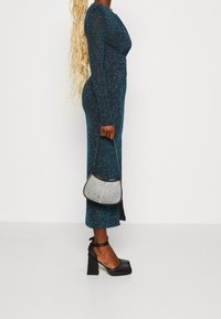 Türkises schimmerndes Kleid mit langen Ärmeln, figurbetontem Design und seitlichem Schlitz; kombiniert mit einer kleinen schwarz-silbernen Handtasche und Pumps mit Blockabsatz.
