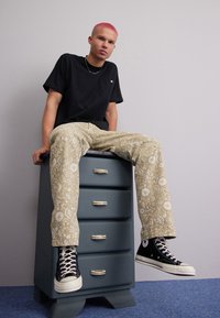 T-shirt nero a maniche corte con taschino, pantaloni beige a fantasia con disegni floreali e sneakers nere alte. Seduto su un comò blu.