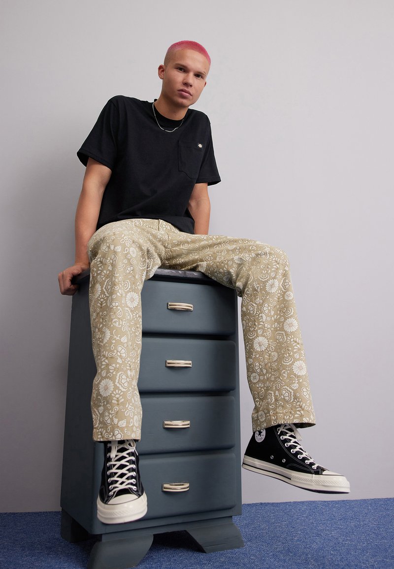 T-shirt nero a maniche corte con taschino, pantaloni beige a fantasia con disegni floreali e sneakers nere alte. Seduto su un comò blu.