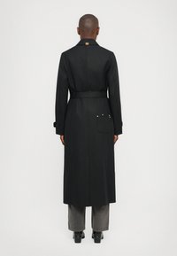 Melna garā mēteli ar jostu, aizmugurējo ventu un dekoratīvām sudraba akcentiem uz kabatas. Pielāgota dizaina, gluda tekstūra un klasiskā apkakle.