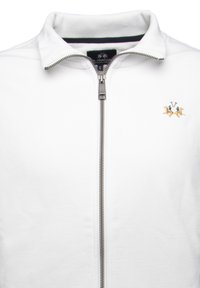 Chaqueta blanca con cremallera alta, que presenta una cremallera plateada y un diseño bordado de dos leones en azul y dorado en el lado izquierdo del pecho.