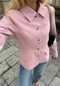 Femme portant un blazer rose boutonné, un jean bleu et des sandales blanches, portant un petit sac noir avec une bandoulière chaîne dorée.