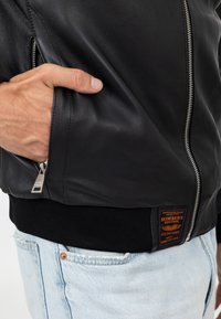Giacca bomber in pelle nera con chiusura zip, polsini a coste neri e un'etichetta arancione distintiva sull'orlo. Sono visibili dei jeans in denim chiaro.