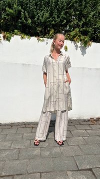 Beige tunika napitnjak s kratkim rukavima i bočnim džepovima, uparen s odgovarajućim širokim hlačama s geometrijskim uzorkom. Crne sandale.