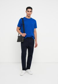 Zils kokvilnas T-krekls ar īsām piedurknēm, neliels logotips uz krūtīm; kombinēts ar tumši ziliem biksēm un baltām sporta apaviem, turēt melnu plecu somu.