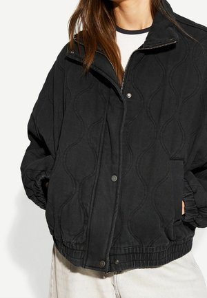 Femme portant une veste matelassée noire avec motifs de coutures ondulées, mains dans les poches, T-shirt blanc en dessous, fond uni et clair.