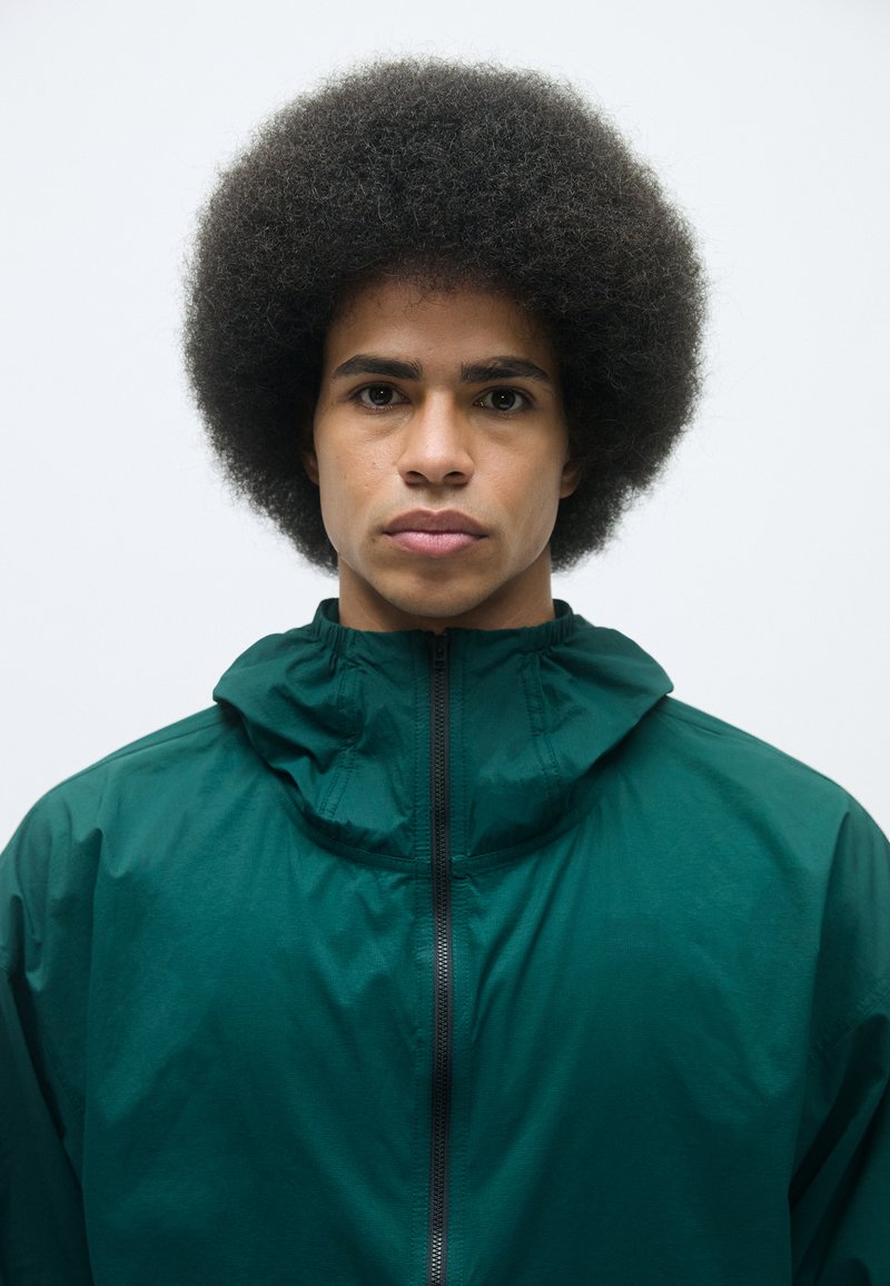Grüne Windjacke mit hohem Kragen und Reißverschluss. Glatte Textur und leichtes Material. Das Model hat eine Afrofrisur.